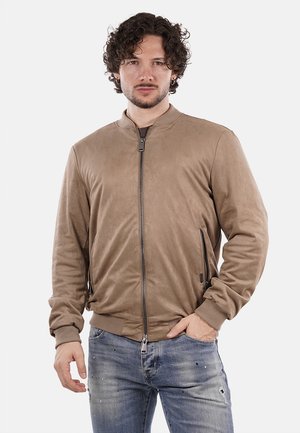 Giacca bomber beige realizzata in materiale liscio, presenta una cerniera frontale, due tasche con zip e polsini a coste. Indossata sopra jeans azzurri.
