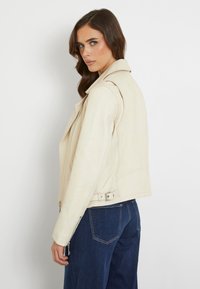 Giacca da biker in pelle beige con dettagli a zip e fibbia, caratterizzata da un design aderente e spalle strutturate. Indossata sopra jeans scuri.