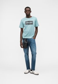 Γαλάζιο βαμβακερό t-shirt με μαύρο γραφικό σχέδιο, συνδυασμένο με μπλε τζιν παντελόνι, μαύρο τσαντάκι μέσης και λευκά αθλητικά παπούτσια με μαύρες ρίγες.