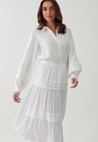 Vestido blanco con volantes y parte superior de camisa abotonada que presenta mangas largas y abullonadas. La tela parece ligera y fluida, con acentos de rayas horizontales.
