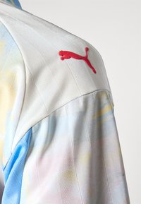 Maglietta sportiva Puma con un design astratto multicolore in azzurro chiaro, giallo e rosa. Presenta un tessuto bianco testurizzato e un accentuazione del logo rosso.