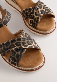 Sandales marron avec des brides croisées à imprimé léopard et une semelle rembourrée, présentées sur une surface claire unie.