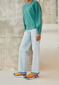 Sudadera verde con puños acanalados, vaqueros rectos azul claro y zapatillas deportivas naranjas con detalles en negro y azul.