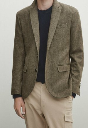 Man wearing a brown textured blazer over a black sweater with beige cargo-style pants, standing against a plain background.