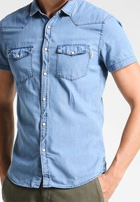 Hombre con una camisa de mezclilla de manga corta azul claro y dos bolsillos en el pecho con botón, de pie con una mano en el bolsillo de pantalones verde oliva.