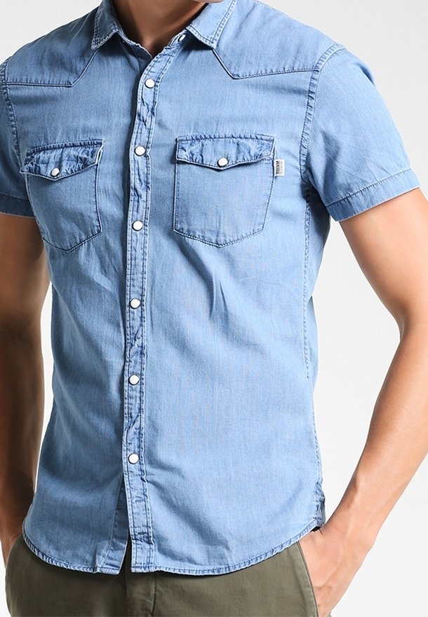 Hombre con una camisa de mezclilla de manga corta azul claro y dos bolsillos en el pecho con botón, de pie con una mano en el bolsillo de pantalones verde oliva.