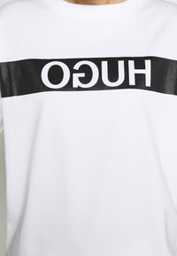 Bílá mikina s černým vodorovným pruhem s nápisem "HUGO" tučnými bílými písmeny, vyrobená z hladké bavlněné směsi.