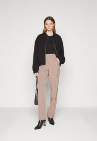 Veste bomber noire texturée sur un haut noir, associée à un pantalon beige taille haute et des bottines noires à talons aiguilles. Sac à main marron.