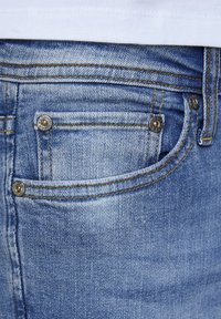 Jeans de mezclilla azul claro con un acabado desgastado, que presentan herrajes de cobre, un bolsillo frontal y costuras amarillas en contraste.