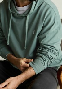 Světle zelená mikina s kapucí a šňůrkami, na hrudi s malým logem. Má měkkou tkaninu a žebrované manžety. Nositá přes bílou košili.