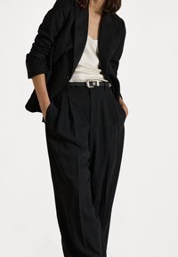 Kvinna i svart kavaj och svarta plisserade byxor med en vit topp och svart bälte, med händerna avslappnat i fickorna mot vit bakgrund.