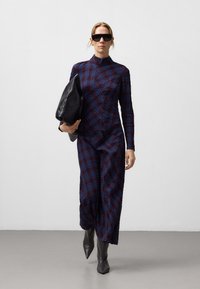 Langärmliges, körperbetontes Kleid in dunklem Karomuster mit Blau- und Burgundtönen. Eine matte schwarze oversized Tasche und kniehohe graue Stiefel runden den Look ab.