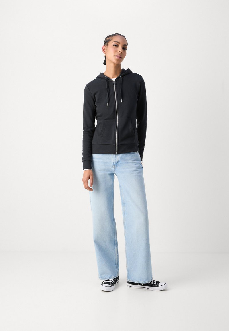 Black zip-up hoodie with drawstrings, two front pockets, paired with light blue wide-leg jeans and black low-top sneakers.