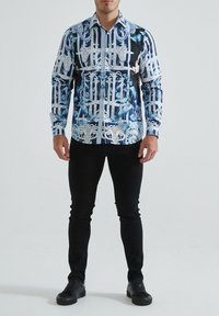 Chemise à manches longues à motifs bleu et blanc, comportant des designs floraux et architecturaux. Associée à un pantalon noir et des baskets.