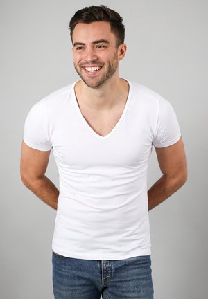 Glunderende man met kort donker haar, die een wit T-shirt met V-hals en blauwe spijkerbroek draagt, staand met handen achter zijn rug.