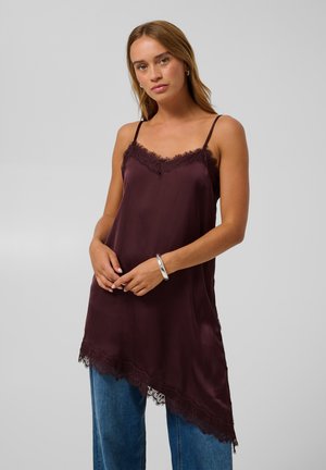 Femme portant un caraco en satin violet foncé avec bordure en dentelle et un jean bleu, debout, les mains jointes devant un fond neutre.