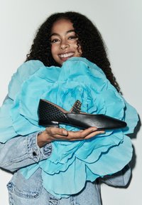 Chaussure en cuir noir avec un bout pointu, présentant un accent en maille noire texturée et une boucle décorative. Tenue devant un grand fond de fleur bleue.
