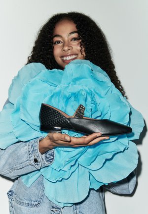 Zapato de cuero negro con puntera afilada, que presenta un acento de malla negra texturizada y una hebilla decorativa. Sostenido frente a un gran fondo de flores azules.