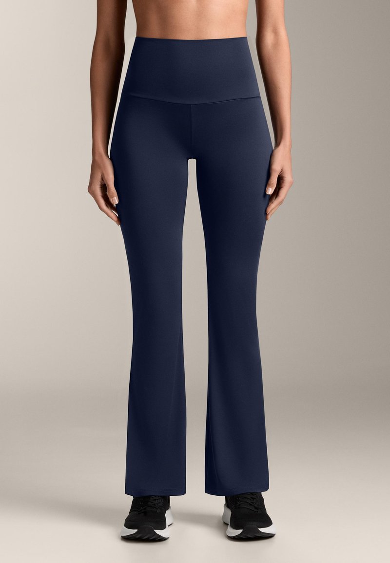 Rozszerzane spodnie do jogi w kolorze granatowym, wykonane z gładkiego, elastycznego materiału. Wysoki stan, w połączeniu z czarnymi butami sportowymi.