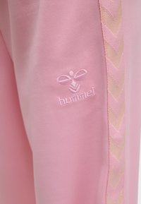 Zbliżenie różowego materiału z haftem logo hummel oraz błyszczącym wzorem w jodełkę na bocznym szwie ubrania.