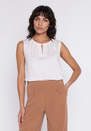 Kobieta o ciemnych włosach do ramion, ubrana w białą, bezrękawnikową bluzkę oraz wysokie brązowe spodnie, stoi z jedną ręką w kieszeni.