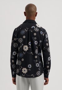 Camisa negra bordada con patrones florales en blanco y azul; mangas largas, con cuello, tela texturizada, cierres de botones, corte holgado.