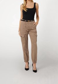 Pantalons cargo marron avec des poignets élastiques, des poches latérales et un devant plat. Portés avec un débardeur noir et des chaussures noires. Ceinture simple incluse.