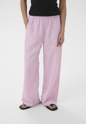 Persona con pantalones sueltos rosa con rayas verticales finas y chanclas negras, de pie con las manos cerca de los bolsillos contra un fondo liso.