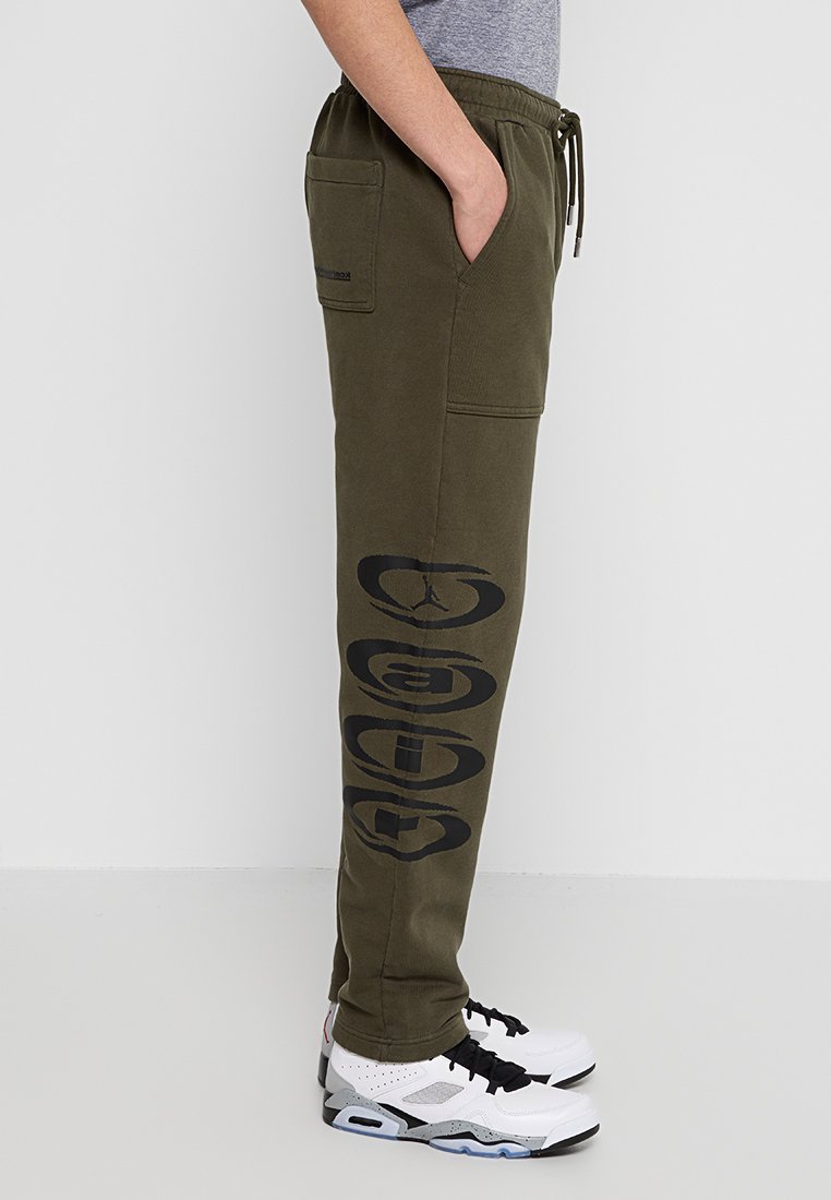 Pantalones de chándal verde olivo en tejido de mezcla de algodón, con bolsillos laterales y un atrevido estampado gráfico negro a lo largo de la pierna izquierda. Cintura con cordón ajustable.