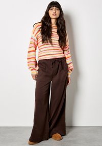 Sweter w wielokolorowe paski z okrągłym dekoltem, połączony z brązowymi spodniami o szerokich nogawkach i beżowymi butami typu slip-on, charakteryzującymi się luźnym krojem.