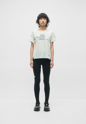 T-shirt léger en tissu évacuant l'humidité de couleur gris clair avec logo, leggings noirs et chaussures de course avec un motif. Le modèle se tient droit devant un fond uni.
