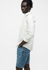 Jeune homme à la peau sombre et aux dreadlocks, portant une chemise blanche à boutons avec les manches retroussées et un short en jean bleu, debout de profil.