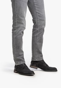 Scarpe in pelle nera a trama con un accento liscio, abbinate a jeans grigi aderenti. Le scarpe presentano una punta arrotondata e una suola bassa.