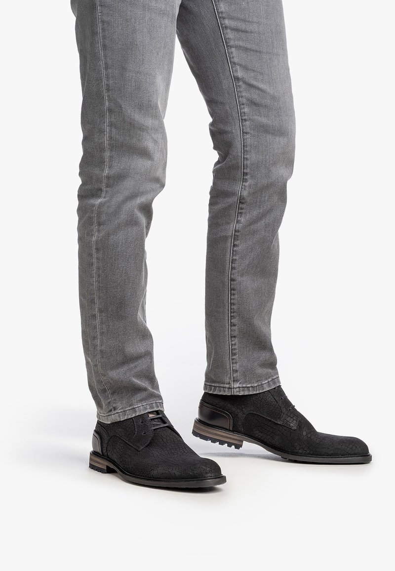 Scarpe in pelle nera a trama con un accento liscio, abbinate a jeans grigi aderenti. Le scarpe presentano una punta arrotondata e una suola bassa.