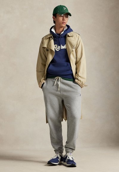 Giovane uomo con occhiali, cappellino verde, trench beige sopra felpa blu navy, pantaloni della tuta grigi e sneaker blu, in piedi con le mani in tasca.