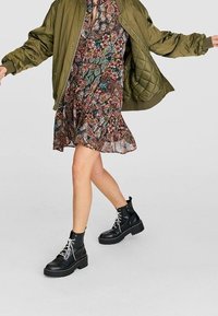 Chaqueta bomber acolchada verde oliva sobre un vestido de colores variados con un dobladillo acampanado. Botines negros de tobillo con cordones blancos y suelas gruesas.