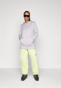 Helllila Kapuzenpullover mit Kängurutasche und Logo, kombiniert mit leuchtend gelben Skihosen und schwarzen Skischuhen. Modernes sportliches Design.