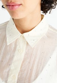 Blusa blanca y transparente con cuello clásico, que presenta dibujos plateados dispersos, dos botones y una tela ligera con textura.