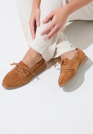 Person, der hviler hænderne på beige linnedbukser og iført brune, ruskindssnøresko på en hvid gulv.