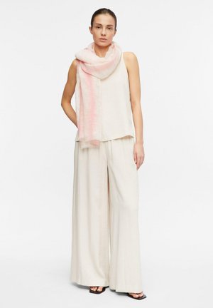 Donna che indossa un top crema senza maniche, pantaloni larghi, sciarpa rosa chiaro e sandali neri con punta aperta, in piedi contro uno sfondo bianco semplice.