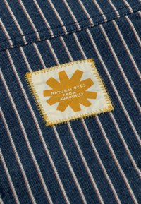 Tissu bleu avec des rayures verticales blanches et rouges, présentant un patch blanc cousu avec un motif de rayon de soleil jaune et le texte "Colorants naturels d'Auroville."