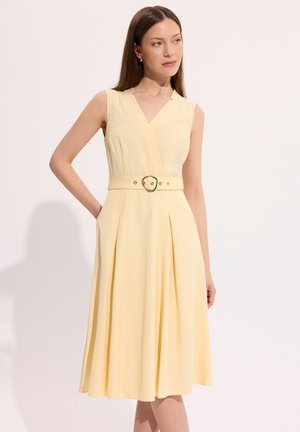 Bez ramiączek, bladożółta sukienka midi z dekoltem w kształcie litery V, plisami i dopasowanym paskiem z okrągłą klamrą noszona przez kobietę z prostymi włosami.