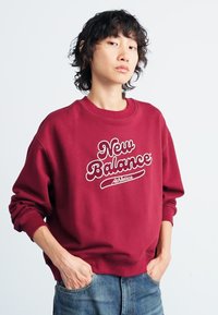 Vynio burgundinės spalvos sportinis megztinis su baltu "New Balance Athletics" tekstu. Minkšta medvilnės medžiaga, apvalus kaklas, laisvas siluetas, įrengtos rankogalio ir apačios dalys.