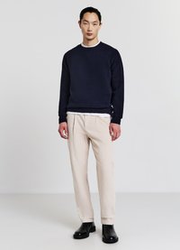 Dunkelblauer Strickpullover mit Rippbündchen, kombiniert mit hellbeigen, plissierten Hosen und schwarzen Stiefeletten, vor einem schlichten Hintergrund stehend.