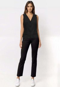 Top nero senza maniche con scollatura a V, caratterizzato da cinque bottoni e tasche laterali, abbinato a pantaloni neri aderenti e sneakers bianche.