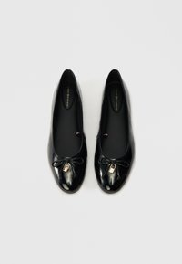 Ballerines en cuir verni noir avec un bout arrondi, détail de nœud noir et une petite touche dorée. Texture lisse et design minimaliste.