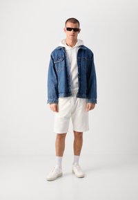 Jeansjakke i blå, hvit hettegenser under, hvite teksturerte shorts, hvite sneakers og svarte solbriller. Enkelt, avslappet antrekk.