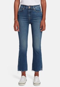 Jeans a vita alta svasati in denim scuro, caratterizzati da orli sfilacciati, due tasche frontali e accenti di cucitura delicata, abbinati a sneakers bianche.