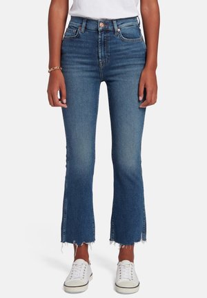 Jeans a vita alta svasati in denim scuro, caratterizzati da orli sfilacciati, due tasche frontali e accenti di cucitura delicata, abbinati a sneakers bianche.