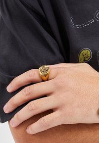 Arany pecsétgyűrű bonyolult gravírozással, sima gyűrűsárral és texturált részletekkel. Kéz elhelyezve egy fekete ing anyagán.